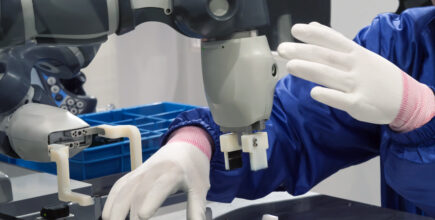 a worker, wearing white fabric gloves, delicately handles a small circuit board. The human work is also being assisted by a collaborative robot.